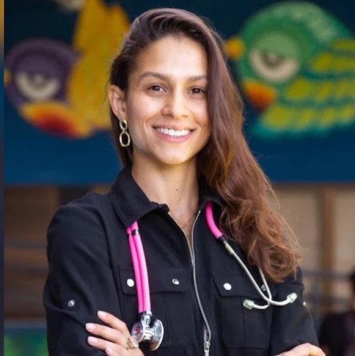 Retrato sorridente da Dra. Elke Louise com um estetoscópio rosa e preto ao redor do pescoço.