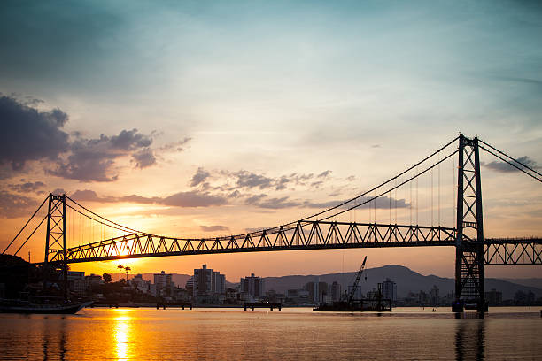 Silhueta da Ponte Hercílio Luz ao pôr do sol sobre a água em Florianópolis.